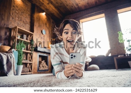 Full size photo of astonished pretty girl laying carpet floor open mouth use smart phone texting daylight flat indoors