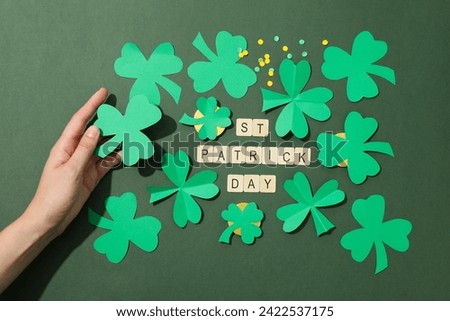 Paper clover leaves in hand, golden coins and letters on green background, top view