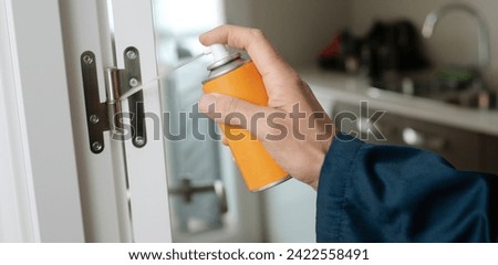 closeup of a caucasian man, in blue overalls, spraying some lubricating oil on the hinge of a white door, in a panoramic format to use as web banner or header Royalty-Free Stock Photo #2422558491