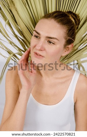Tall slender European young woman with blonde hair in a bun in a white singlet, against the background of a palm leaf on a white background