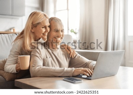 Retirement and internet fun concept. Excited smiling senior spouses using modern laptop computer, websurfing or watching film online, resting on sofa at home, embracing, drinking coffee, free space Royalty-Free Stock Photo #2422625351