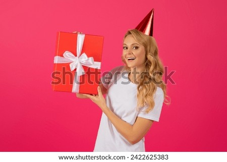 Happy young blonde woman wearing festive birthday hat and holding wrapped gift box, smiling to camera showing bday surprise present standing on pink studio background. Fun party, holiday