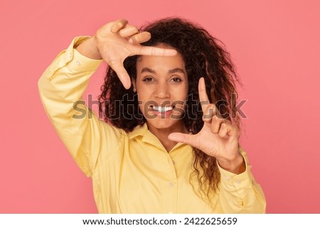 Imaginative black woman making frame with her hands, smiling, wearing yellow blouse, symbolizing artistic vision on pink background