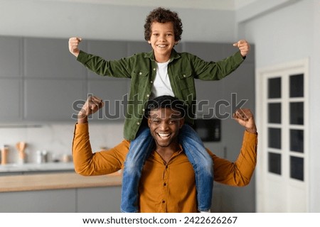 Black preteen boy on his dad's shoulders triumphantly raises his fists in the air, as they both flex their biceps, displaying strength and bonding at home Royalty-Free Stock Photo #2422626267