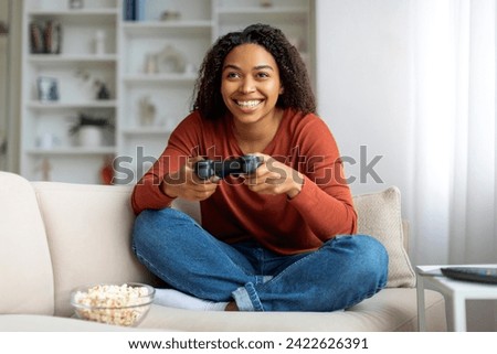 Domestic Fun. Cheerful Young Black Woman Playing Video Games At Home, Happy African American Lady Using Joystick And Smiling While Sitting On Couch With Popcorn In Living Room, Free Space