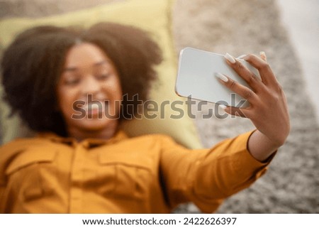 Smiling millennial African American woman with curly hair takes a selfie while lying down, capturing a joyful moment with her smartphone in a cozy home environment, blurred