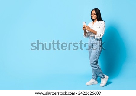 Indian Lady In Casual Using Smartphone Texting Standing On Blue Background, Studio Shot. Young Middle Eastern Woman Websurfing Via Gadget. Great Mobile App Concept. Full Length, Empty Space Royalty-Free Stock Photo #2422626609