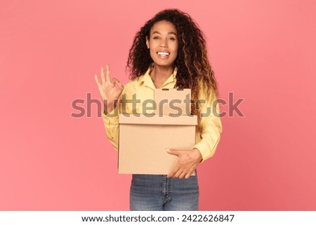 Confident young black woman, with warm smile, holds box parcels and makes an okay gesture against pink background, expressing satisfaction, positivity and approval