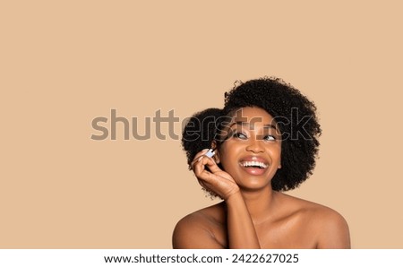 Cheerful young black woman with curly hair applying mascara, looking upwards at free space with joyful expression, against sofa beige studio background