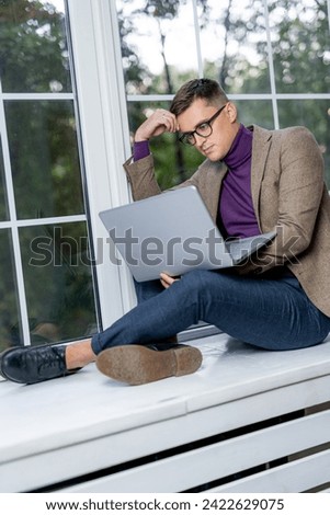 Young man sitting on the windowsill and working on laptop