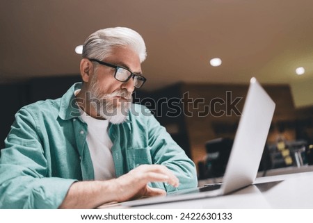 Focused senior Caucasian man with a beard working intently on a laptop in an indoor setting, wearing glasses and a green shirt, symbolizing professional dedication and digital engagement in later life Royalty-Free Stock Photo #2422630133