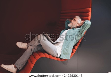 Senior Caucasian businessman relaxing in modern office lounge, reflecting on work with laptop computer on lap, symbolizing work-life balance and strategic thinking in a comfortable work environment