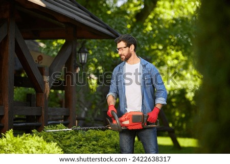 Skilled landscaper in gloves shaping evergreen hedge with trimmer at summer day. Front view of happy man taking care of conifer bush with electric lopper, against wooden gazebo. Concept of gardening. Royalty-Free Stock Photo #2422638317