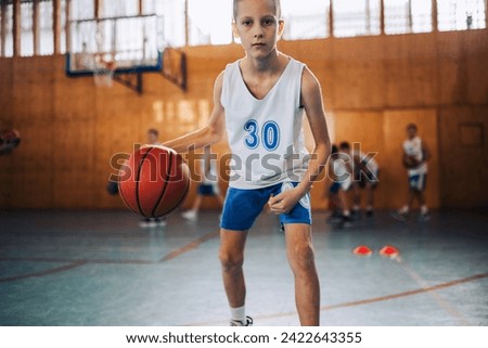 A young basketball kid is dribbling a ball on court and having training. A dynamic junior athlete in action dribbling a ball and practicing basketball at indoor court. In blurry background is his team