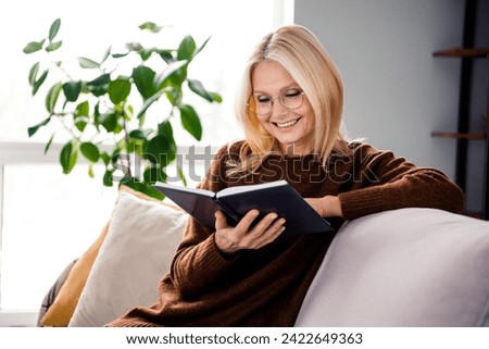 Photo of cheerful lovely cute woman pensioner sitting on couch reading detective novel indoors Royalty-Free Stock Photo #2422649363