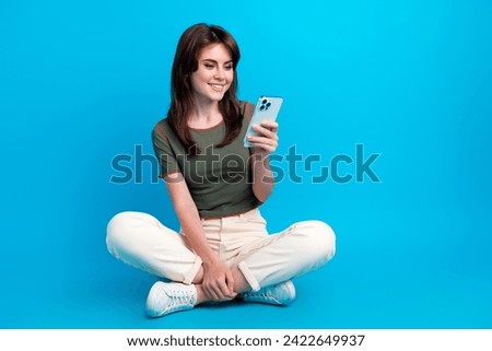 Full size photo of optimistic woman wear green t-shirt sit on floor look at smartphone read email isolated on blue color background