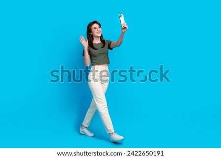 Full size profile portrait of friendly girl walk hold smart phone arm wave make selfie walk empty space isolated on blue color background