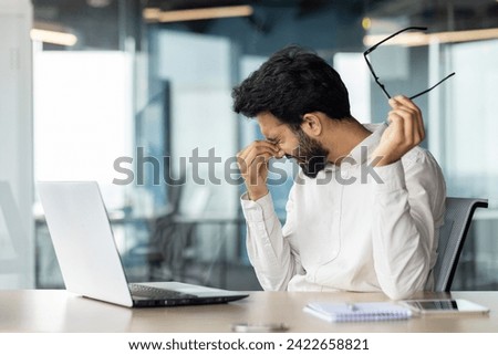Stressed Indian man at an office desk feeling exhausted and overtired from work, holding his glasses and massaging his nose bridge. Royalty-Free Stock Photo #2422658821