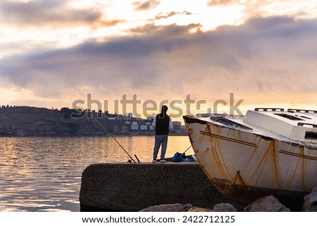 A male fisherman stands on the seashore in the port next to a boat at sunset and fishes with a fishing rod against the background of the sky and horizon