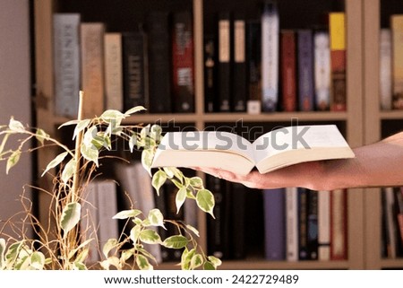 an open book held in one hand, next to a table with a bookcase in the background. Royalty-Free Stock Photo #2422729489