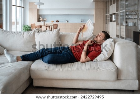 Overheated perspiring man lying on sofa, waving with hand fan, suffering from high temperature in room because of broken conditioner, absence of ventilator, air blower. Male feeling unwell, dehydrated Royalty-Free Stock Photo #2422729495
