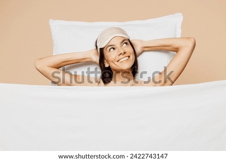 Young minded Latin woman wears pyjamas jam sleep eye mask rest relax at home under duvet be lost in reverie mood look aside isolated on plain pastel light beige background studio. Night nap concept