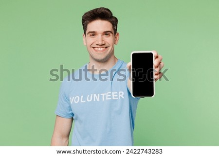 Young man wear blue t-shirt white title volunteer hold use showing blank screen mobile cell phone isolated on plain pastel green background. Voluntary free work assistance help charity grace concept