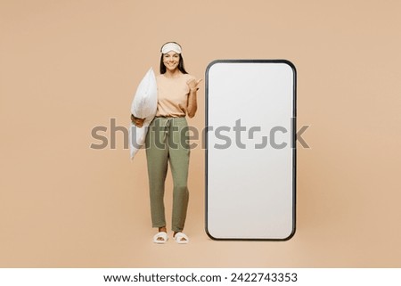Full body young Latin woman wear pyjamas jam sleep eye mask rest relax point on big huge blank screen area mobile cell phone isolated on plain pastel beige background. Good mood night home nap concept Royalty-Free Stock Photo #2422743353