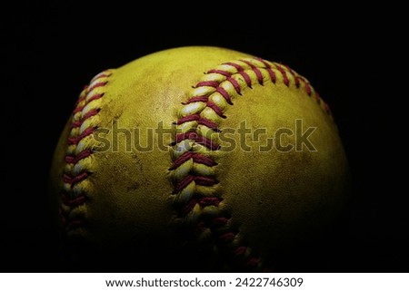 A yellow softball with red seams on a black background.
