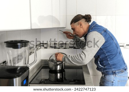 Caucasian man cooking in the kitchen with a pot and a ladle by hand in the natural light, on a ceramic hob, is tasting food, side view. Concept of male doing housework