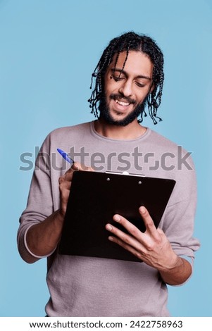 Smiling arab man holding clipboard and writing plan checklist with cheerful facial expression. Carefree young person wearing casual clothes taking notes and filling application