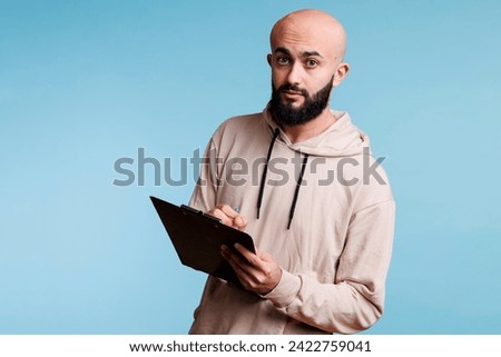 Arab man taking notes on clipboard and looking at camera. Young bald arabian person wearing casual hoodie holding checklist, planning and writing with pen studio portrait