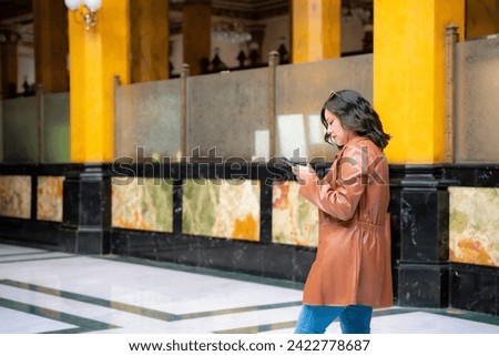 Happy young mexican woman tourist in embroidered top looking around while standing in Postal palace in downtown Mexico City and in light against blurred interior