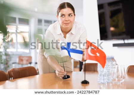 Positive young woman putting little flag of Finland on the table with flag of Turkey in conference room for international negotiations