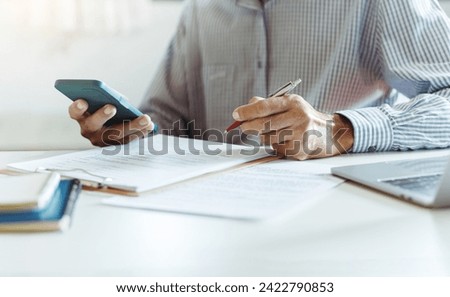 Asian businessman holding mobile phone reviewing document reports at office workplace with laptop. legal expert, professional lawyer reading and checking financial documents or insurance contract
