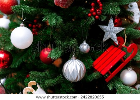 A vibrant Christmas tree adorned with white and red ornaments, glittery silver star, and a whimsical red sled, set against a backdrop of dark green fir branches.