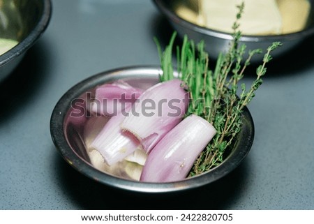 Raw shallots alongside fresh thyme and chives in a metal bowl, prepared for culinary use in a kitchen setting.
