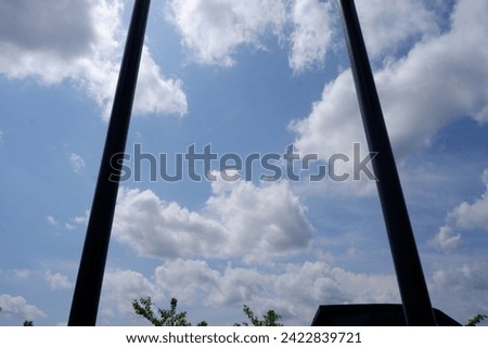 A beautiful photo of a clear and serene sky filled with white clouds, framed by two dark vertical poles and silhouettes of a roof and tree tops. This photo is suitable for themes such as nature.