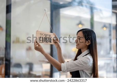 Asian female entrepreneur turning the 'Sorry We Are Closed' sign with a content smile after a day's work at her cafe.
