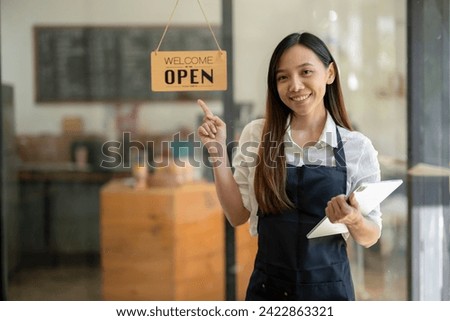 A friendly Asian shop owner smiles and points to a 'Welcome Open' sign, holding a tablet at the entrance of her cafe.

