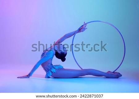 Flexible, talented teen girl, rhythmic gymnast in sparkling bodysuit performing with hoop against gradient studio background in neon light. Concept of sport, grace, competition, art, youth, hobby