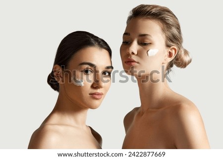 Organic skin care products promotion. Beautiful girls with well-kept skin standing with cream on cheeks against white studio background. Concept of natural beauty, cosmetology and cosmetics, skincare Royalty-Free Stock Photo #2422877669