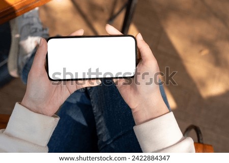 A close-up shot features a woman watching videos on her smartphone while relaxing on a bench outdoors, showcasing a white-screen mockup. Royalty-Free Stock Photo #2422884347