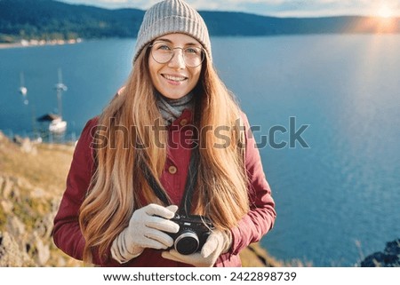 Portrait of a smiling hipster woman in glasses and autumn outfit, holding photo camera, standing on the cliff with a scenic lake view. Solo traveler, explorer, freelance photographer concept