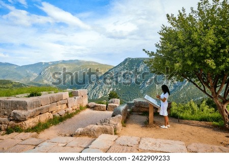 Journeys into Greek history:  A long-haired woman, wearing white dress  studying history from board at Apollo Temple ruins, illuminated by sunset. Delphi, Greece.