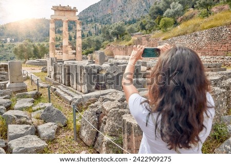 A long-haired woman in a white dress photographing the ancient temple complex of Athena Pronaia in Delphi. Sunny day, blue sky. Archaeological site, UNESCO World Heritage Site, Delphi, Greece.