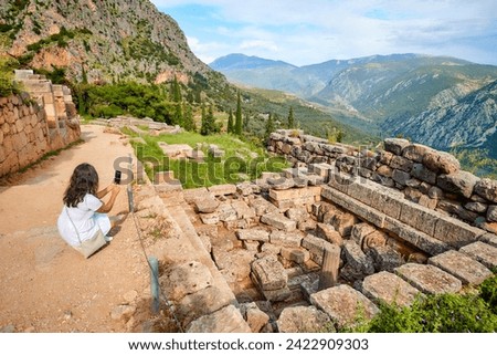 Journeys into Greek history:  A long-haired woman, wearing white dress  taking pictures of the Apollo Temple ruins, illuminated by sunset. Delphi, Greece.