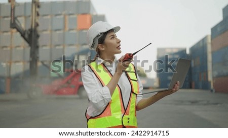 Asian engineer woman in safety uniform talking on walkie-talkie and woking on laptop for control loading containers box from cargo freight ship for import export. Logistic, Transportation concept Royalty-Free Stock Photo #2422914715