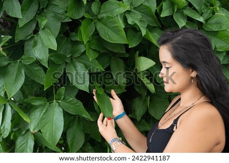 Portrait of beautiful young woman with black hair looking to the side touching the green leaves on a wall. Person traveling.