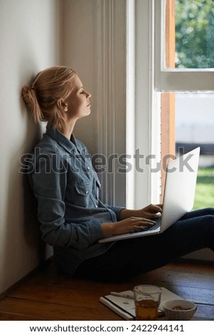 Woman, freelancer and laptop by window, writer and internet connection on floor for info. Female person, editor and website for research or online news, blogging and typing an article on technology